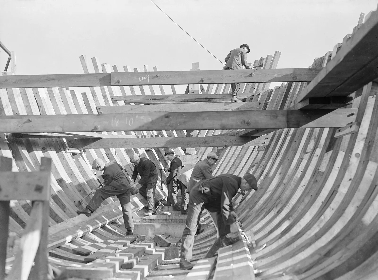 Shipwrights work inside a wooden hull under construction. The frames rise from the keel along the floor.